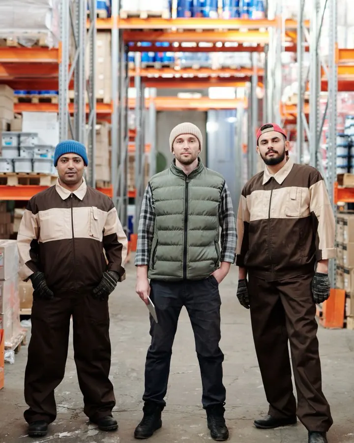 Trois employés d'entrepôt en uniforme, debout entre les rayonnages de stockage.